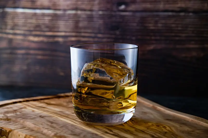 Close-up of whiskey glass with ice cube on wooden surface, moody lighting