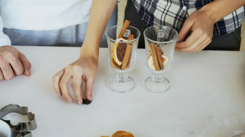 Hands arranging cinnamon sticks and orange slices in glasses on a table