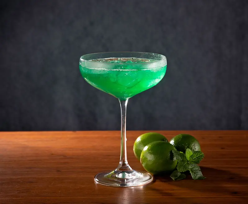 A refreshing green cocktail served in a coupe glass, garnished with fresh limes