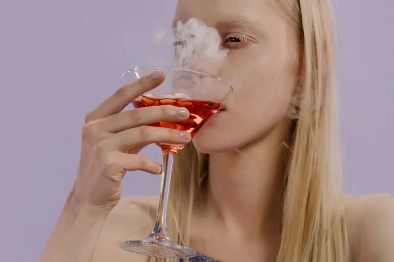 A woman savoring a vibrant, smoky cocktail in a martini glass with blonde hair indoors